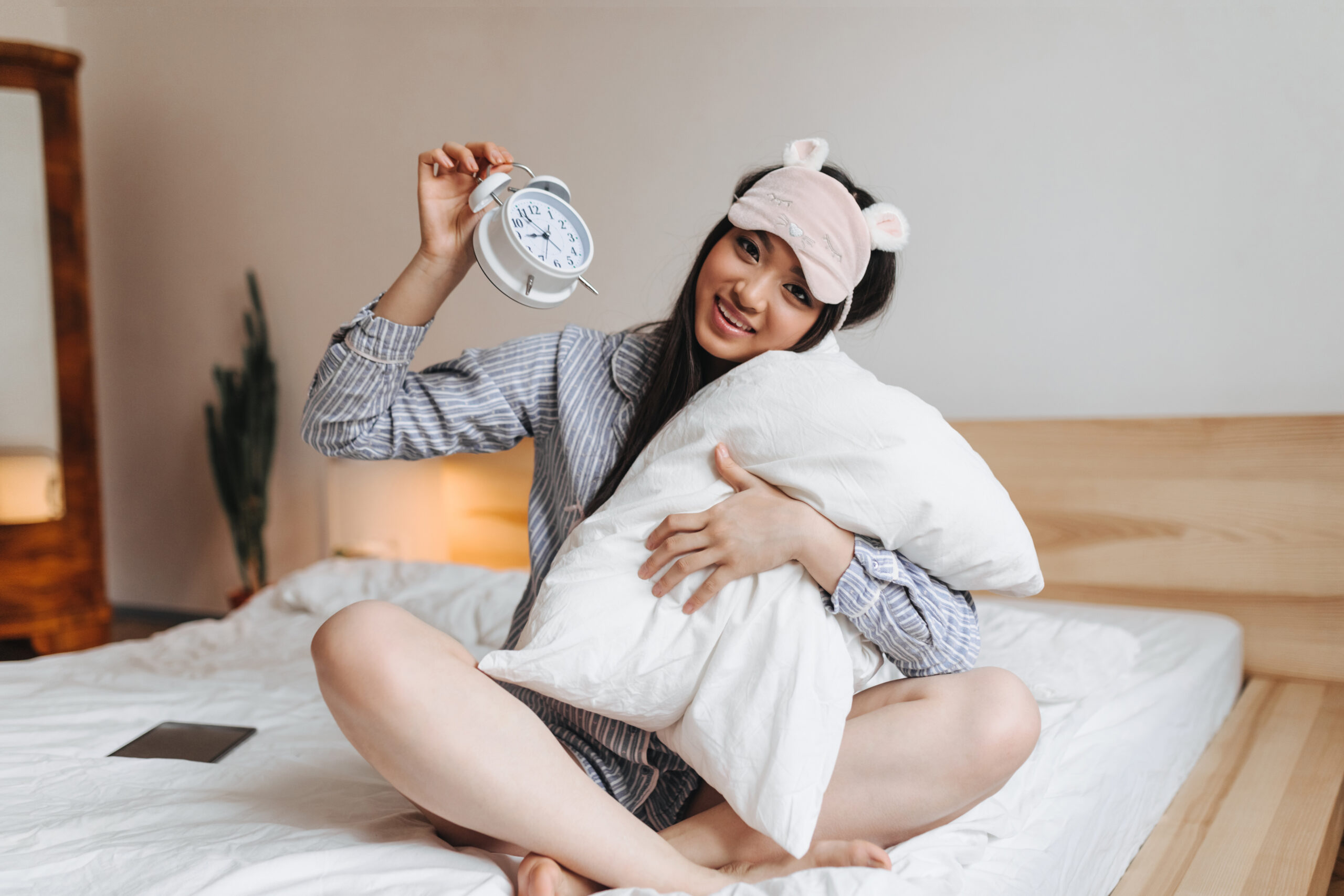 Girl sits on bed and hugs pillow. Woman in pajamas holding white alarm clock and smiling.