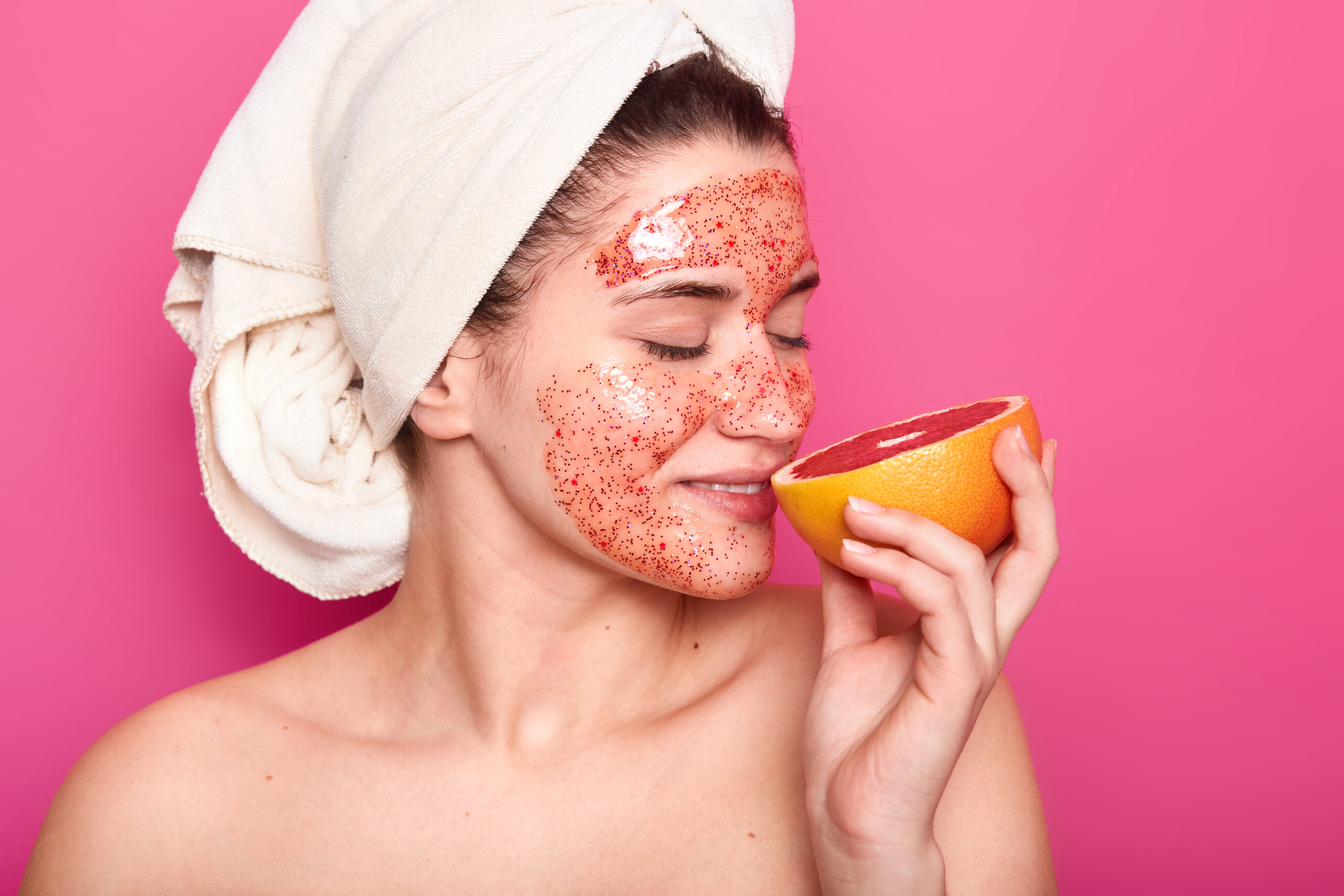 Studio shot of pleasant looking young European woman smelling grapefruit, has white towel on head, showing her naked shoulders, posing isolated over pink background. Beauty and skin care concept.